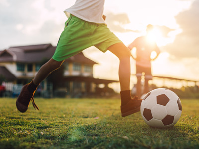 Ein Kind, das auf einem Fußballfeld einen Fußball tritt. Im Hintergrund sind Personen und Häuser sichtbar.